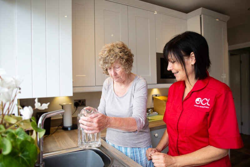 Older lady filling up a jug with tap water, supported by  female care worker with black shoulder length hair and wearing a red ECL tunic.